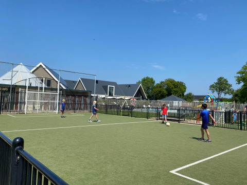 Kinderen voetballen op een veldje bij een glampingverblijf, met huisjes en speeltuin op de achtergrond.