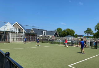 Kinderen voetballen op een veldje bij een glampingverblijf, met huisjes en speeltuin op de achtergrond.