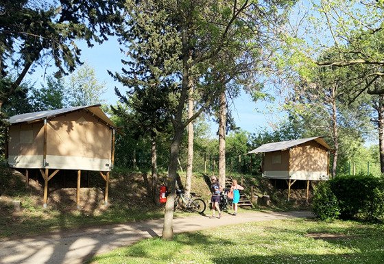Twee verhoogde Bivouac Comfort safaritenten op een groene camping met fietsers op de voorgrond.
