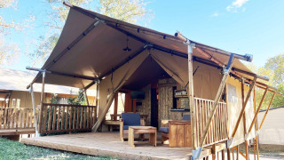 Glamping-accommodatie met tentstructuur, houten veranda en zitplaatsen buiten in een natuurlijke omgeving.