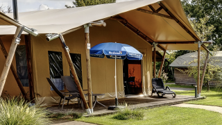 Outdoor photo of a luxury safari tent lodge with deck chairs and umbrella on a grassy campsite setting