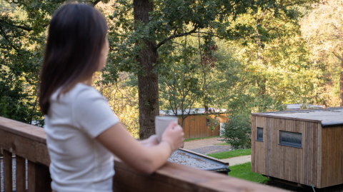 Frau mit Tasse blickt von Balkon auf Holzchalets im Ferienpark Hameau de la Semois, Belgien.
