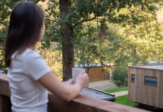 Woman with a mug stands on a balcony overlooking cabins and trees at Hameau de la Semois, Belgium.