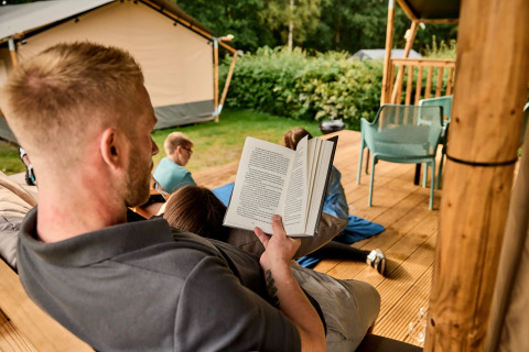Mand læser en bog på verandaen af en safariteltsuite med børn der leger på Vestbirk Camping, Danmark.