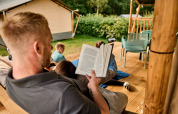 Uomo legge un libro sulla veranda di una tenda safari con bambini che giocano a Vestbirk Camping, Danimarca.