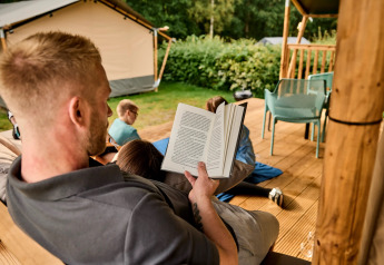 Homme lisant un livre sur la terrasse d'une tente safari, enfants jouant, à Vestbirk Camping, Danemark.