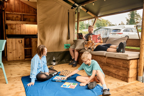 Familie hygger sig i en safaritelt med eget bad på Vestbirk Camping, børn leger spil på et tæppe.