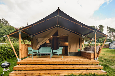 Tente safari avec sanitaires, terrasse avec chaises et canapé, entourée de verdure et d'arbres.
