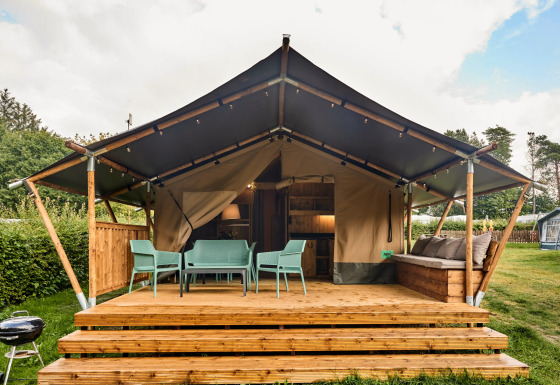 Safari-Zelt mit Sanitäranlagen, Terrasse mit Stühlen und Sofa, umgeben von Grün und Bäumen.