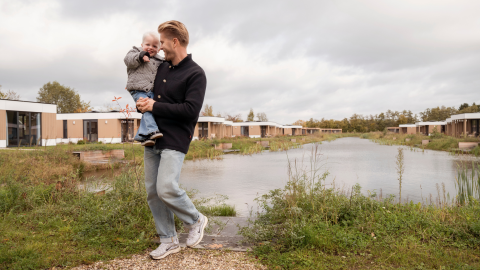 Man holding child by the lake at Water lodge 6 Deluxe in Hofparken Wiltershaar, Netherlands, cloudy day.