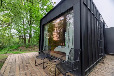 Cabaña Nokken Forest negra con ventana grande, dos sillas y mesa pequeña en terraza de madera rodeada de bosque.