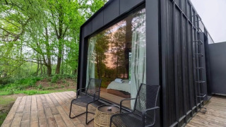 Schwarze Nokken Forest Cabin mit großen Fenstern, zwei Stühlen und Tisch auf Holzdeck im grünen Wald.
