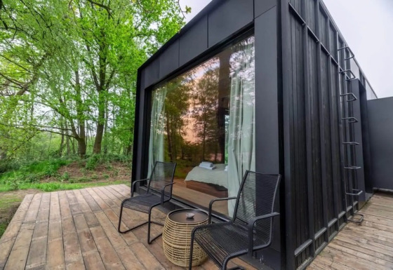 Zwarte Nokken Forest Cabin met groot raam, twee stoelen en tafeltje op houten terras midden in het bos.