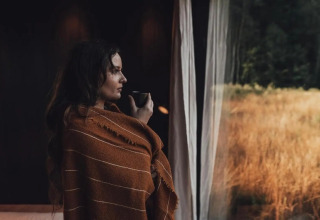 Mujer con manta y taza contempla el campo desde Nokken Forest Cabin en Wilsumer Berge, Alemania.