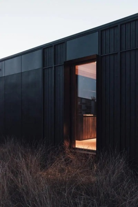 Cabaña negra Nokken Forest en Wilsumer Berge, Alemania, con ventana abierta y césped alto en el exterior.