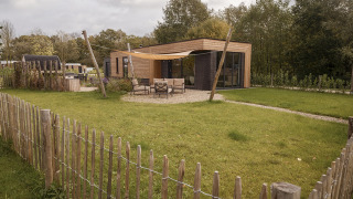 Foto einer modernen Holzhütte mit Terrasse, Gartenmöbeln und eingezäuntem Garten in grüner Umgebung.