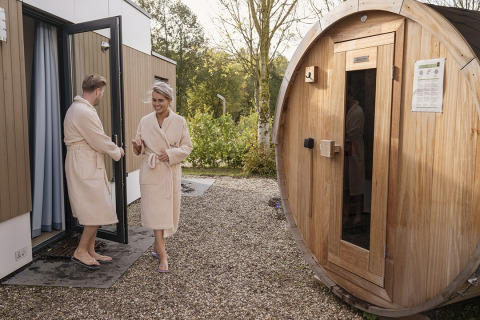 Couple in bathrobes steps out of a lodge and walks towards a wooden outdoor barrel sauna.