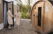 Pareja con batas sale de una cabaña y camina hacia una sauna de madera al aire libre.