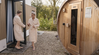 Ein Paar im Bademantel verlässt eine Hütte und geht zu einer runden Holzsauna im Freien.
