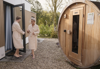 Un couple en peignoir sort d'un lodge et se dirige vers un sauna rond en bois à l'extérieur.
