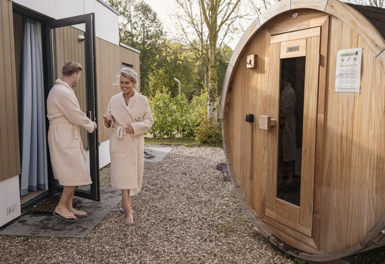 Couple in bathrobes steps out of a lodge and walks towards a wooden outdoor barrel sauna.
