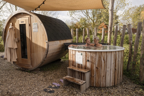 Un couple se détend dans un bain à remous en bois près d’un sauna tonneau dans un lodge confortable.