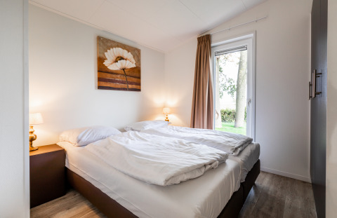 Lichte slaapkamer met tweepersoonsbed, witte lakens, lampen, schilderij en uitzicht op de tuin.