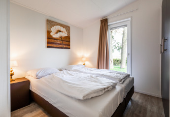 Dormitorio luminoso con cama doble, ropa de cama blanca, lámparas, cuadro y vista al jardín.