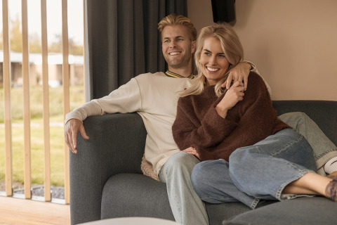 Een lachend koppel ontspant samen op een sofa in een lodge, zonlicht schijnt door de ramen binnen.