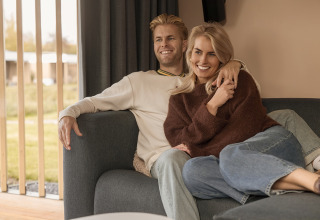 Una pareja sonriente se relaja en un sofá de una cabaña, con luz natural entrando por las ventanas cercanas.