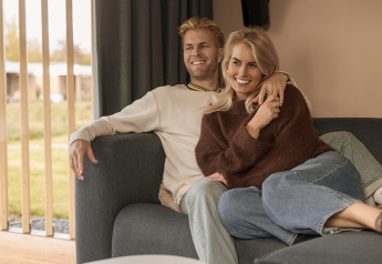 Ein lächelndes Paar entspannt gemeinsam auf einem Sofa in einer Lodge, Tageslicht strömt herein.