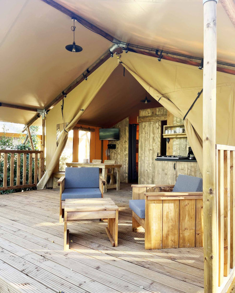 Interno di tenda safari con veranda in legno, poltrone, tavolo e piccola cucina all'aperto.