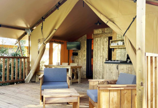 Interno di tenda safari con veranda in legno, poltrone, tavolo e piccola cucina all'aperto.