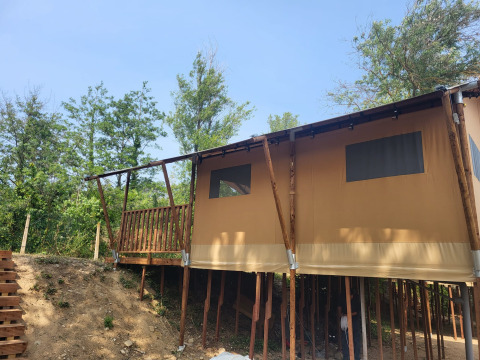 Tenda safari su palafitte con terrazza in legno in mezzo al verde, sotto un cielo limpido e azzurro.