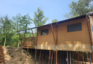 Tenda safari su palafitte con terrazza in legno in mezzo al verde, sotto un cielo limpido e azzurro.