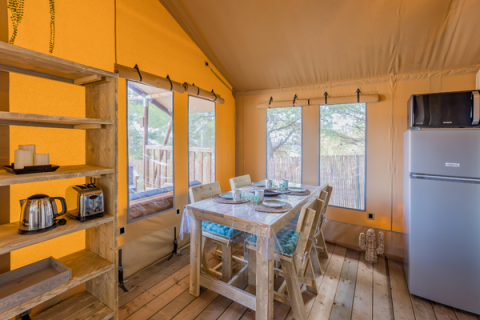 Interno di una tenda safari con tavolo da pranzo, sedie, scaffali, frigorifero e grandi finestre sulla natura.