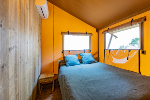 Interno di una tenda safari con letto matrimoniale, cuscini blu, arredi in legno e ampie finestre.