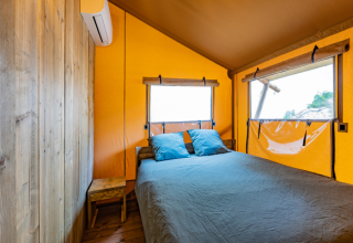 Interno di una tenda safari con letto matrimoniale, cuscini blu, arredi in legno e ampie finestre.