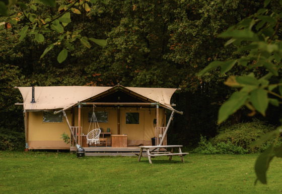 Tente safari Glamping tent La Vida sur une pelouse verte, entourée d’arbres, avec terrasse et banc.