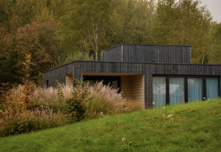 Moderne Villa aus dunklem Holz im Resort de Parel, Niederlande, umgeben von grünem Gras und Wald.