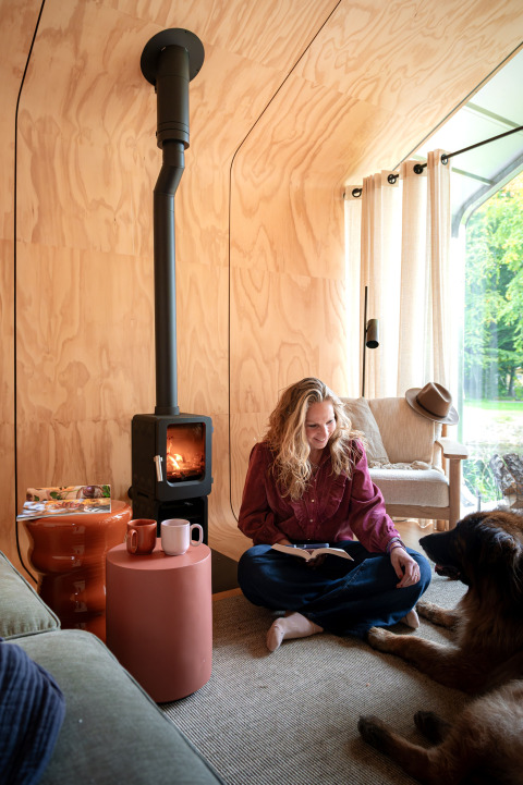 Femme lisant devant un poêle dans le lodge Wrap house à Resort de Parel, Pays-Bas, avec un chien.