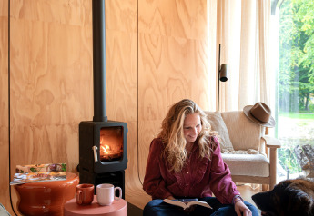 Frau liest am Kamin im Wrap House Lodge bei Resort de Parel, Niederlande, mit Hund neben sich.