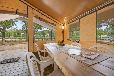 Eethoek van Safaritent Harmony met houten tafel, witte stoelen en zicht op het kampeerterrein door ramen.