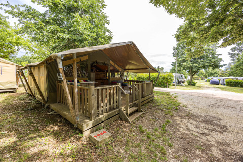 Tienda safari Safaritent Relax con porche de madera, rodeada de árboles y campamento en el fondo.
