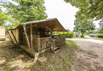 Tenda safari Safaritent Relax con veranda in legno, circondata da alberi e un campeggio sullo sfondo.