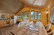 Interior de la Safaritent Harmony con mesa de madera, sillas, cocina y vista a la naturaleza exterior.