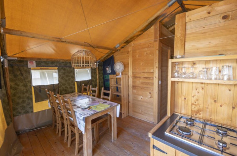 Interno di tenda safari con tavolo da pranzo, sedie, fornello a gas e decorazioni in legno.