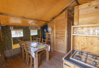 Interno di tenda safari con tavolo da pranzo, sedie, fornello a gas e decorazioni in legno.