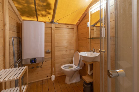 Baño en tienda safari con paredes de madera, inodoro, lavabo, ducha, calentador y suelo de madera.