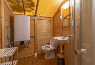Bathroom inside a safari tent with wooden walls, toilet, sink, shower, water heater, and wood floor.